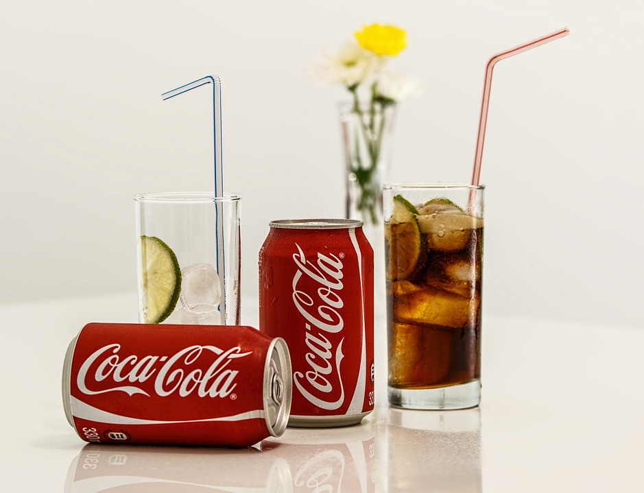A photo of two cans of coca cola, two glasses with ice - one is full with coca cola, the other contains ony ice and a lime slice.