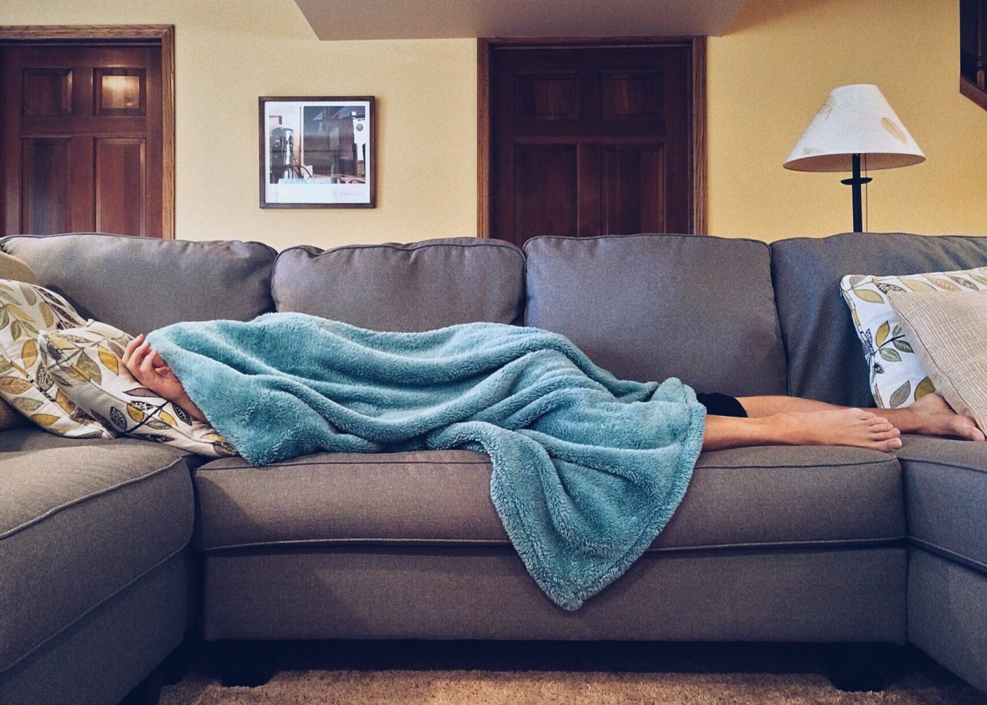 A woman lies on a grey sofa, covered by a blue blanket. Only her feet are showing.