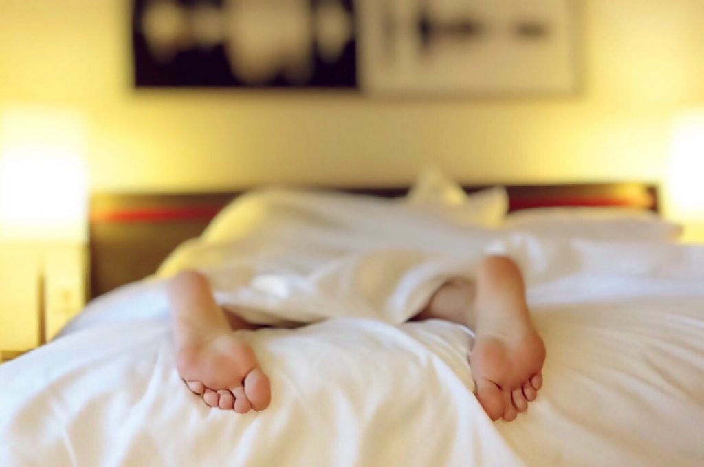 A woman lies on a bed, only her feet can be seen poking out from under a white sheet.