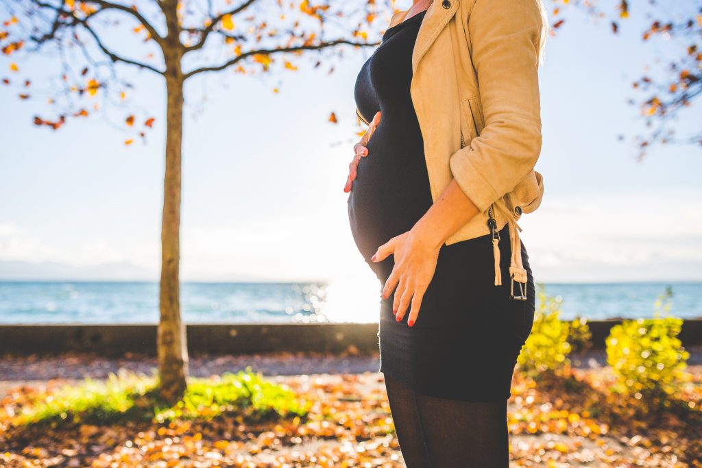 A pregnant woman in a black dress and tan jacket stands under some trees on the water front.