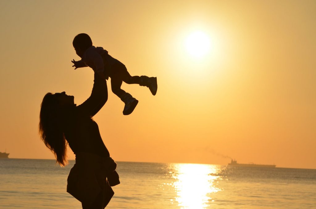 A silhouette of a mother holding her baby up in the air against a waterfront sunset.