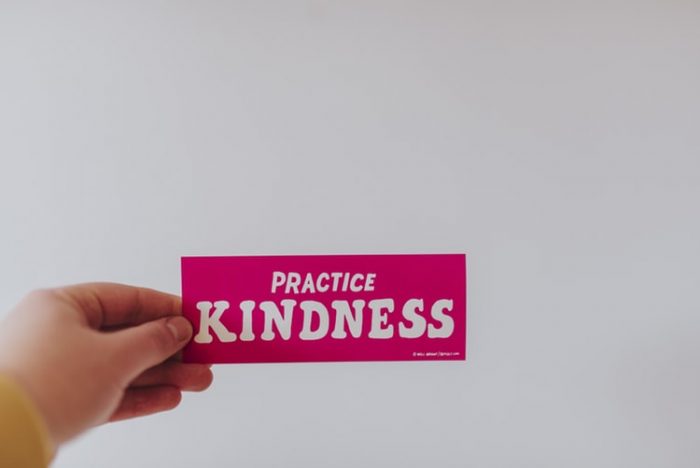 A note is held up against a white wall. It reads "practice kindness" on red paper.
