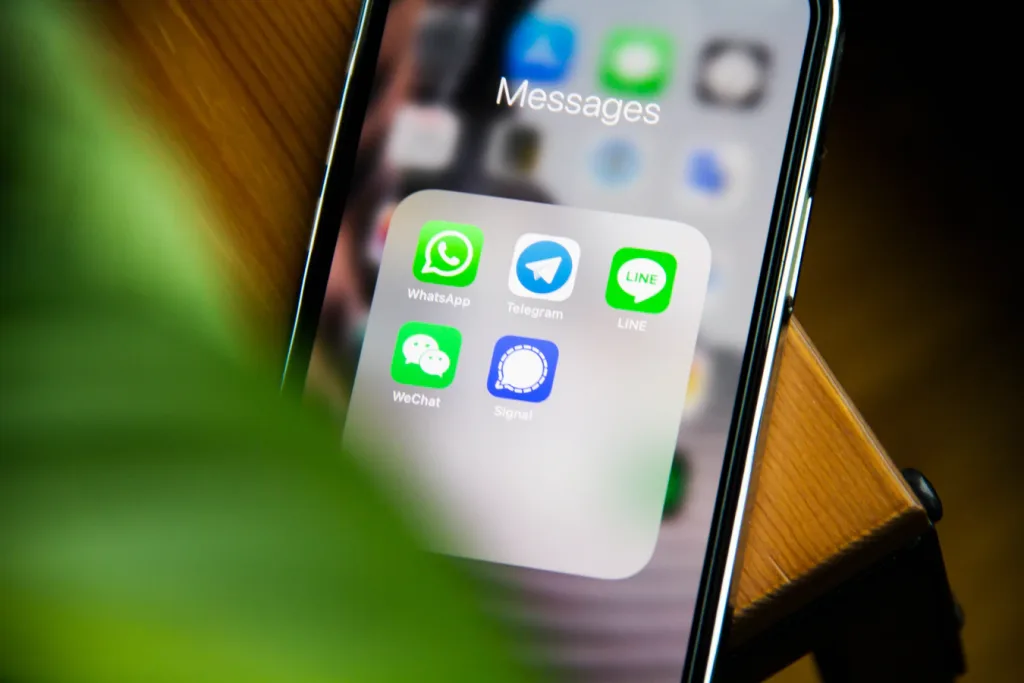 A colour photograph of a phone screen on a table with a blurred plant in the foreground. One the screen of the iPhone the messages folder is open displaying apps which can send various message forms.